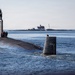 A large military submarine is navigating through open coastal waters with several crew members visible on the forward deck near the sail (conning tower). The submarine's stern section displays markings including 'PROJ' and numerical depth/draft indicators (2-7). In the background, a small island with buildings and a flagpole is visible, along with a lighthouse further in the distance.