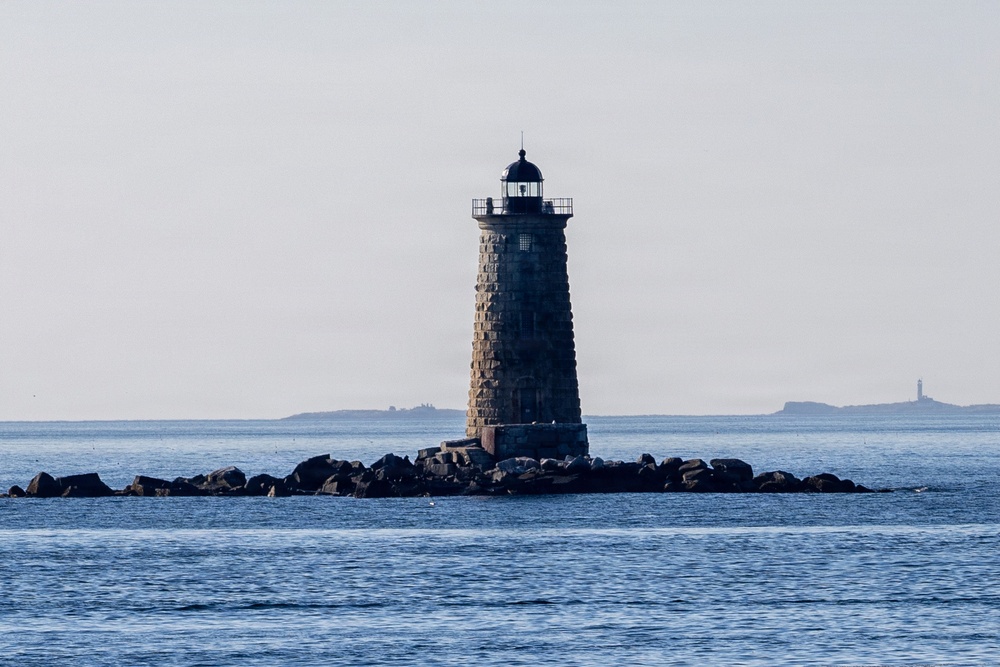 A solitary granite lighthouse tower stands on a small rocky outcropping surrounded by open ocean water. The tower has a distinctive tapered cylindrical form constructed of rough-cut stone blocks, topped with a black lantern room and railing. In the distant background, at least one additional lighthouse or structure is faintly visible on the horizon.