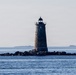 A solitary granite lighthouse tower stands on a small rocky outcropping surrounded by open ocean water. The tower has a distinctive tapered cylindrical form constructed of rough-cut stone blocks, topped with a black lantern room and railing. In the distant background, at least one additional lighthouse or structure is faintly visible on the horizon.
