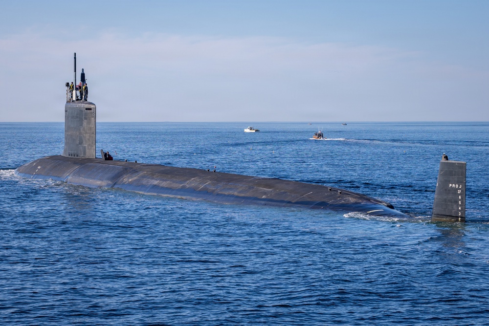 A large military submarine is navigating on the surface of open ocean water under clear skies. Several personnel are visible on the sail (conning tower), and two smaller escort vessels are visible in the background. A vertical fin or rudder structure with markings is visible at the stern of the submarine.
