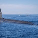 A large military submarine is navigating on the surface of open ocean water under clear skies. Several personnel are visible on the sail (conning tower), and two smaller escort vessels are visible in the background. A vertical fin or rudder structure with markings is visible at the stern of the submarine.