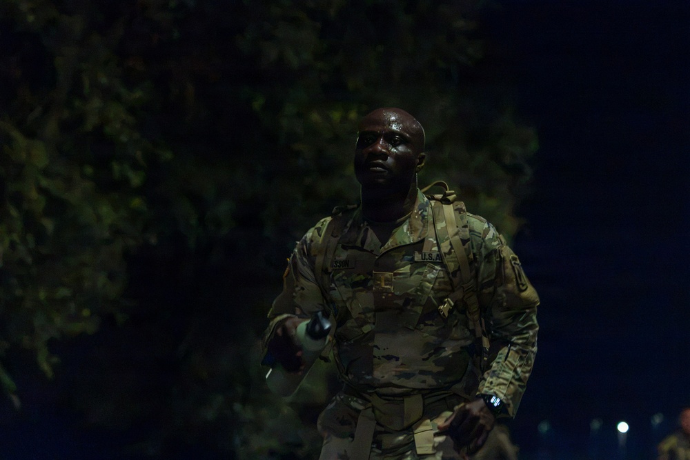 A U.S. Army soldier in OCP (Operational Camouflage Pattern) uniform is photographed at night, appearing to be engaged in a ruck march or physical training exercise. The soldier is visibly sweating and carrying a rucksack along with what appears to be a water bottle or flashlight. The 'U.S. Army' tape is clearly visible on the chest of the uniform.