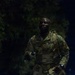 A U.S. Army soldier in OCP (Operational Camouflage Pattern) uniform is photographed at night, appearing to be engaged in a ruck march or physical training exercise. The soldier is visibly sweating and carrying a rucksack along with what appears to be a water bottle or flashlight. The 'U.S. Army' tape is clearly visible on the chest of the uniform.