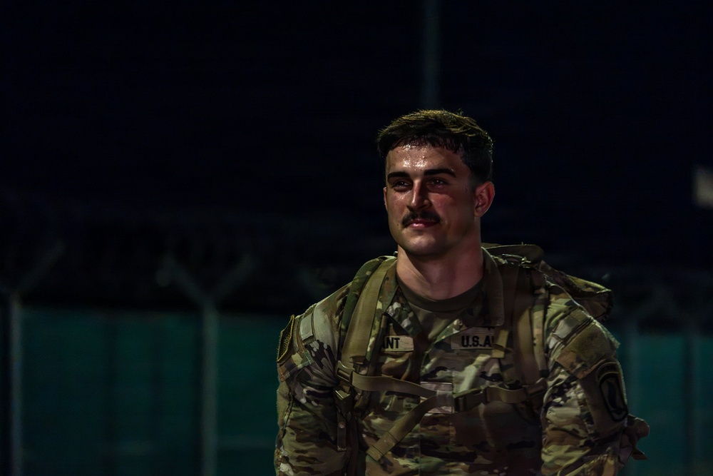 A U.S. Army soldier is photographed at night wearing MultiCam (OCP) camouflage uniform with a tactical vest and rucksack. The soldier appears to be sweating heavily, suggesting recent physical exertion, and is looking off to one side. The image is dramatically lit with a single light source against a dark background.
