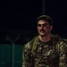 A U.S. Army soldier is photographed at night wearing MultiCam (OCP) camouflage uniform with a tactical vest and rucksack. The soldier appears to be sweating heavily, suggesting recent physical exertion, and is looking off to one side. The image is dramatically lit with a single light source against a dark background.