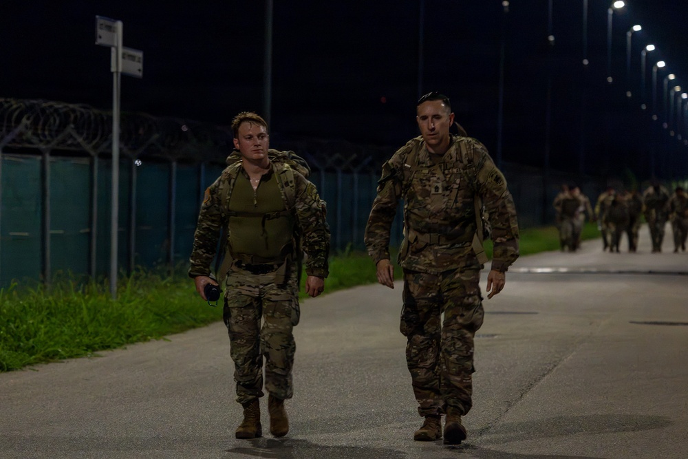 Two military personnel in camouflage uniforms are walking along a paved road at night, with additional soldiers visible in the background. The scene appears to be a ruck march or physical training exercise conducted on a military installation. The area is illuminated by street lights and bordered by a security fence with razor wire.