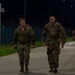 Two military personnel in camouflage uniforms are walking along a paved road at night, with additional soldiers visible in the background. The scene appears to be a ruck march or physical training exercise conducted on a military installation. The area is illuminated by street lights and bordered by a security fence with razor wire.