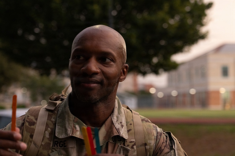 A male soldier in US Army OCP (Operational Camouflage Pattern) uniform is photographed in a candid outdoor setting, smiling and looking off to the side. He is holding what appear to be colored popsicles or freeze pops in his hand. The background shows trees and blurred buildings consistent with a military installation.