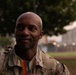 A male soldier in US Army OCP (Operational Camouflage Pattern) uniform is photographed in a candid outdoor setting, smiling and looking off to the side. He is holding what appear to be colored popsicles or freeze pops in his hand. The background shows trees and blurred buildings consistent with a military installation.