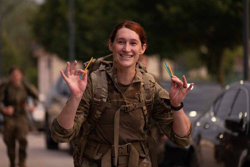 A female soldier in MultiCam/MTP camouflage uniform is smiling broadly at the camera while holding small colored sticks or markers in both hands. She is wearing a tactical backpack and appears to be outdoors on a street or base road. The mood is cheerful and celebratory, suggesting a positive moment during a military exercise or event.