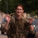 A female soldier in MultiCam/MTP camouflage uniform is smiling broadly at the camera while holding small colored sticks or markers in both hands. She is wearing a tactical backpack and appears to be outdoors on a street or base road. The mood is cheerful and celebratory, suggesting a positive moment during a military exercise or event.