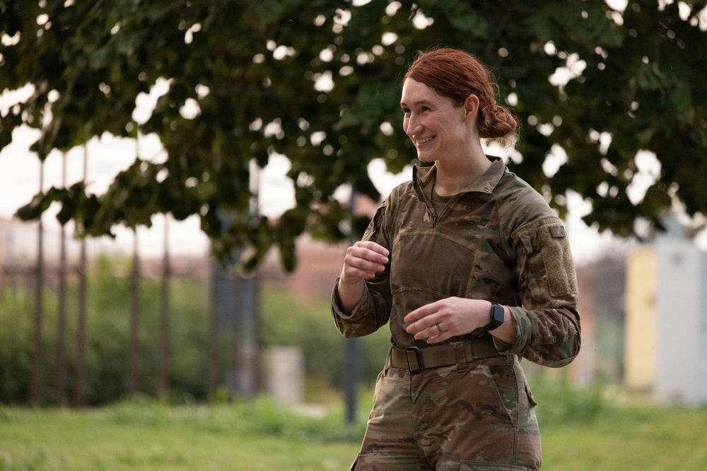 A female military service member wearing MultiCam/OCP camouflage uniform is photographed outdoors in what appears to be a relaxed, candid moment. She has red hair pulled back, is smiling, and appears to be in motion or conversation. The background shows trees, grass, and blurred structures consistent with a military installation or base.