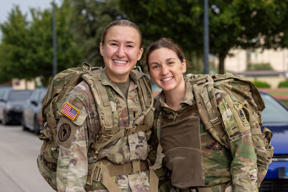 Two female U.S. Army soldiers in Operational Camouflage Pattern (OCP) uniforms are posing together outdoors, smiling broadly at the camera. Both are wearing full tactical rucksacks and gear, suggesting they have completed or are participating in a ruck march or field exercise. The setting appears to be a military installation based on the background infrastructure.
