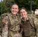 Two female U.S. Army soldiers in Operational Camouflage Pattern (OCP) uniforms are posing together outdoors, smiling broadly at the camera. Both are wearing full tactical rucksacks and gear, suggesting they have completed or are participating in a ruck march or field exercise. The setting appears to be a military installation based on the background infrastructure.