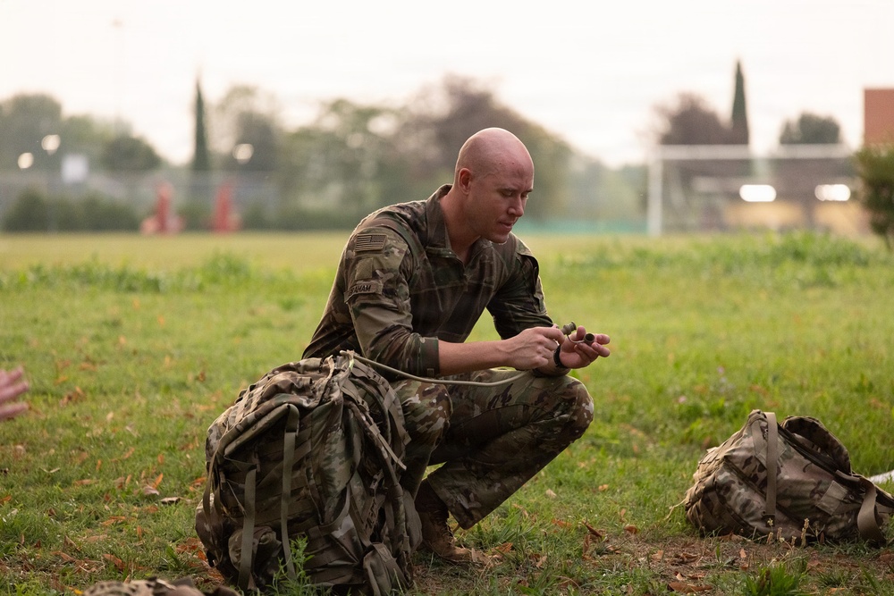 A bald male soldier in MultiCam/OCP camouflage uniform is crouched on a grass field, closely inspecting or manipulating a small object connected to a cord or strap. A large military rucksack is positioned beside him, and another pack is visible in the background to the right. The setting appears to be an open field or sports ground during early morning or overcast conditions.