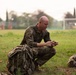 A bald male soldier in MultiCam/OCP camouflage uniform is crouched on a grass field, closely inspecting or manipulating a small object connected to a cord or strap. A large military rucksack is positioned beside him, and another pack is visible in the background to the right. The setting appears to be an open field or sports ground during early morning or overcast conditions.