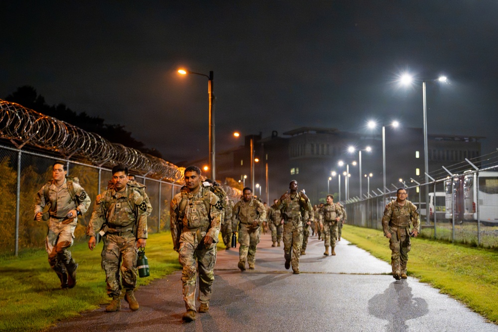 A group of military personnel in camouflage uniforms are conducting a night ruck march or physical training exercise along a paved path. The scene is illuminated by street lights and takes place within a secured military installation, identifiable by the chain-link fence topped with concertina/razor wire. The soldiers are carrying rucksacks and appear to be moving at a brisk pace.