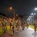 A group of military personnel in camouflage uniforms are conducting a night ruck march or physical training exercise along a paved path. The scene is illuminated by street lights and takes place within a secured military installation, identifiable by the chain-link fence topped with concertina/razor wire. The soldiers are carrying rucksacks and appear to be moving at a brisk pace.
