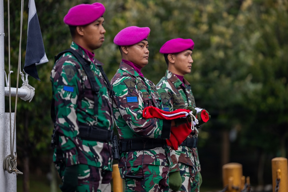 Three military personnel in camouflage uniforms and distinctive magenta/pink berets stand at attention during what appears to be a formal ceremony. The center soldier is holding a folded red and white flag, suggesting a flag-raising or ceremonial parade. Their uniforms and beret color are consistent with the Indonesian Marine Corps (Korps Marinir TNI-AL).