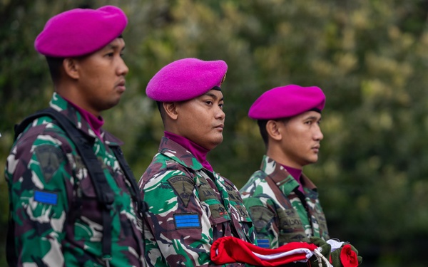 Three military personnel in camouflage uniforms and distinctive magenta/pink berets stand at attention during what appears to be a formal ceremony. The center soldier is holding a folded red and white flag, suggesting a flag-raising or ceremonial parade. Their uniforms and beret color are consistent with the Indonesian Marine Corps (Korps Marinir TNI-AL).