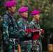 Three military personnel in camouflage uniforms and distinctive magenta/pink berets stand at attention during what appears to be a formal ceremony. The center soldier is holding a folded red and white flag, suggesting a flag-raising or ceremonial parade. Their uniforms and beret color are consistent with the Indonesian Marine Corps (Korps Marinir TNI-AL).