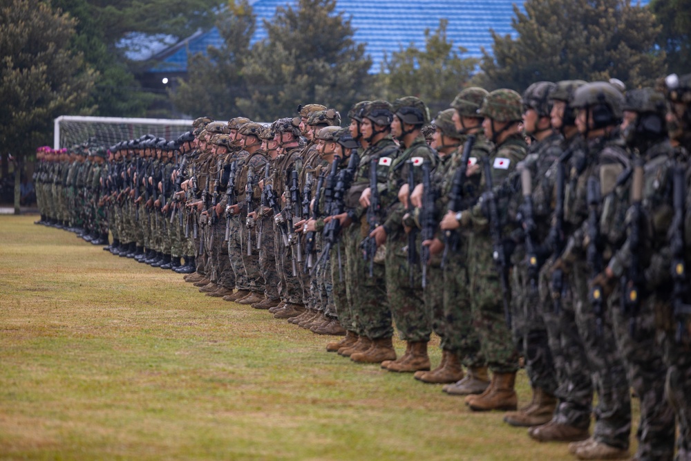 A large formation of military personnel standing in a long line on a grass field, appearing to be a joint military exercise or ceremony. Soldiers wearing Japanese Ground Self-Defense Force uniforms with Japanese flag patches are visible alongside what appear to be U.S. Marines in different camouflage patterns. All personnel are armed and in full combat gear.