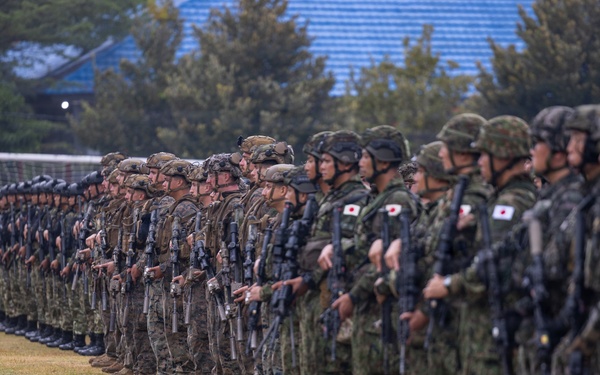 A large formation of military personnel standing in a long line on a grass field, appearing to be a joint military exercise or ceremony. Soldiers wearing Japanese Ground Self-Defense Force uniforms with Japanese flag patches are visible alongside what appear to be U.S. Marines in different camouflage patterns. All personnel are armed and in full combat gear.