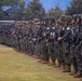 A large formation of military personnel standing in a long line on a grass field, appearing to be a joint military exercise or ceremony. Soldiers wearing Japanese Ground Self-Defense Force uniforms with Japanese flag patches are visible alongside what appear to be U.S. Marines in different camouflage patterns. All personnel are armed and in full combat gear.