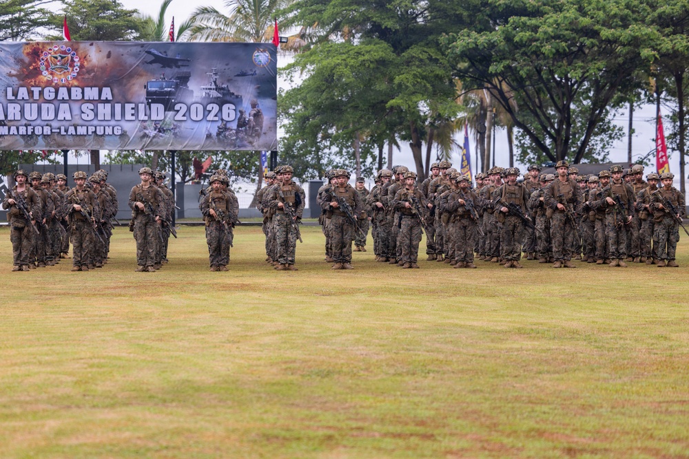 A large formation of armed military personnel in camouflage uniforms stands on a grass field during what appears to be an opening or closing ceremony for 'Garuda Shield 2026,' a joint military exercise held in Lampung, Indonesia. The soldiers are equipped with rifles and full combat gear, arranged in organized rows facing the camera. A large banner in the background identifies the exercise as 'LATGABMA GARUDA SHIELD 2026 MARFOR-LAMPUNG.'