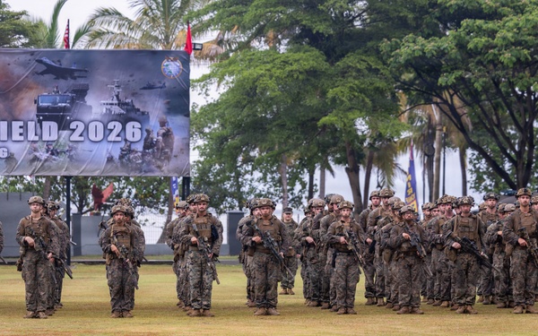 A large formation of armed military personnel in camouflage uniforms stands on a grass field during what appears to be an opening or closing ceremony for 'Garuda Shield 2026,' a joint military exercise held in Lampung, Indonesia. The soldiers are equipped with rifles and full combat gear, arranged in organized rows facing the camera. A large banner in the background identifies the exercise as 'LATGABMA GARUDA SHIELD 2026 MARFOR-LAMPUNG.'