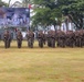 A large formation of armed military personnel in camouflage uniforms stands on a grass field during what appears to be an opening or closing ceremony for 'Garuda Shield 2026,' a joint military exercise held in Lampung, Indonesia. The soldiers are equipped with rifles and full combat gear, arranged in organized rows facing the camera. A large banner in the background identifies the exercise as 'LATGABMA GARUDA SHIELD 2026 MARFOR-LAMPUNG.'