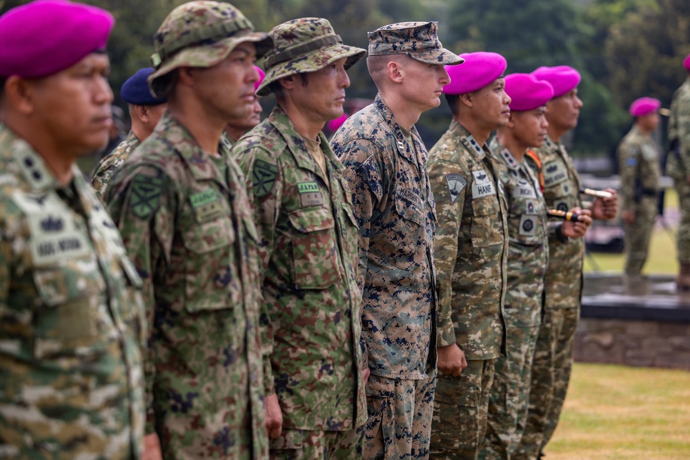 A multinational military formation shows soldiers from multiple nations standing at attention in a formal ceremony or parade. Visible uniform patches and headgear indicate participation from at least Japan, the United States (Marine Corps), and other allied nations. The soldiers are aligned in a single row on a grass field, suggesting a joint military exercise or official ceremony.