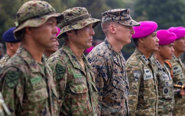 A multinational military formation shows soldiers from multiple nations standing at attention in a formal ceremony or parade. Visible uniform patches and headgear indicate participation from at least Japan, the United States (Marine Corps), and other allied nations. The soldiers are aligned in a single row on a grass field, suggesting a joint military exercise or official ceremony.