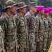A multinational military formation shows soldiers from multiple nations standing at attention in a formal ceremony or parade. Visible uniform patches and headgear indicate participation from at least Japan, the United States (Marine Corps), and other allied nations. The soldiers are aligned in a single row on a grass field, suggesting a joint military exercise or official ceremony.