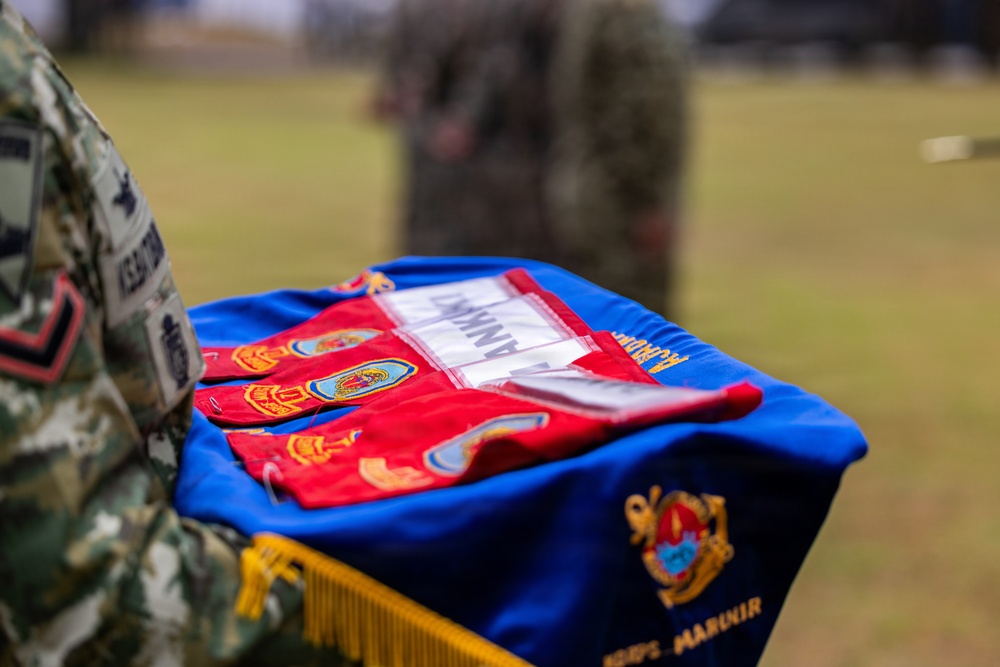 A military ceremony is taking place outdoors, with a soldier in camouflage uniform holding a blue ceremonial cushion or tray displaying red guidon flags with unit patches and emblems. The blue fabric bears gold embroidery including what appears to be a unit crest and partial text reading 'MARUOIR' or similar. The setting appears to be a formal military event such as a change of command or unit activation ceremony.