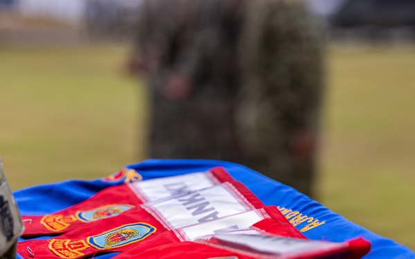 A military ceremony is taking place outdoors, with a soldier in camouflage uniform holding a blue ceremonial cushion or tray displaying red guidon flags with unit patches and emblems. The blue fabric bears gold embroidery including what appears to be a unit crest and partial text reading 'MARUOIR' or similar. The setting appears to be a formal military event such as a change of command or unit activation ceremony.