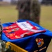 A military ceremony is taking place outdoors, with a soldier in camouflage uniform holding a blue ceremonial cushion or tray displaying red guidon flags with unit patches and emblems. The blue fabric bears gold embroidery including what appears to be a unit crest and partial text reading 'MARUOIR' or similar. The setting appears to be a formal military event such as a change of command or unit activation ceremony.