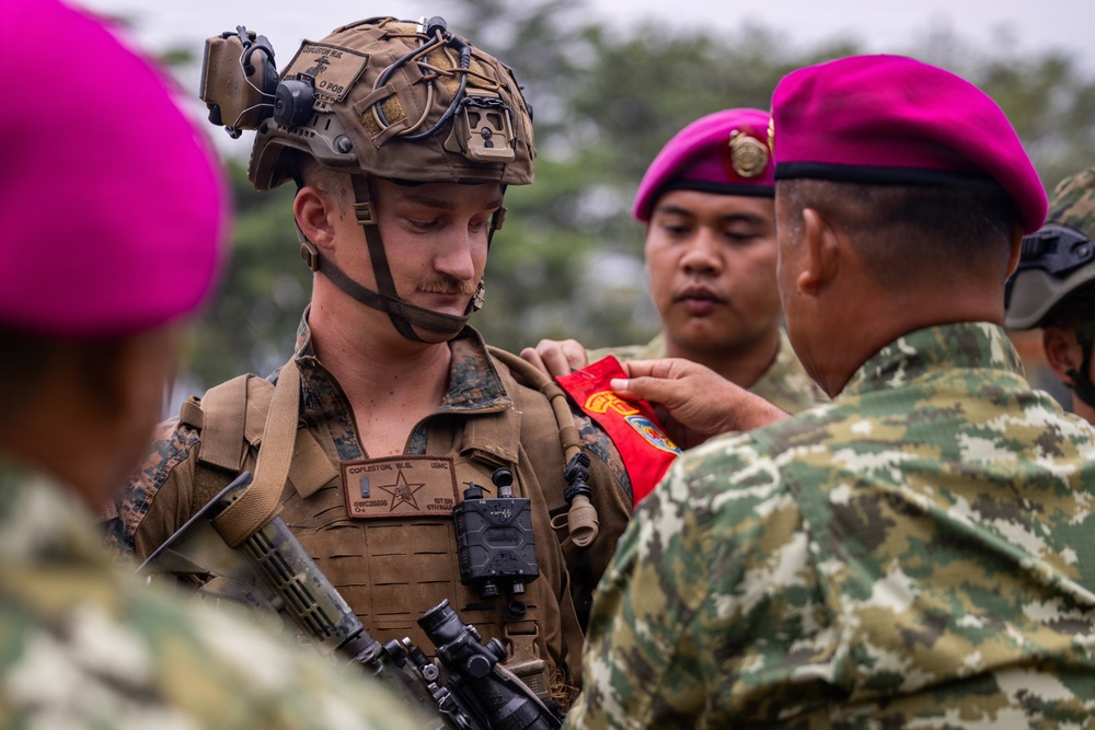 A U.S. Marine in full combat gear is receiving what appears to be a patch or insignia from a foreign military soldier wearing a distinctive magenta/pink beret. The scene suggests a joint military exercise or ceremony involving U.S. Marines and what appears to be Indonesian Marine Corps (Korps Marinir) personnel, identifiable by their characteristic magenta berets and camouflage uniforms. This type of exchange is common during bilateral military training exercises.