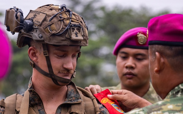 A U.S. Marine in full combat gear is receiving what appears to be a patch or insignia from a foreign military soldier wearing a distinctive magenta/pink beret. The scene suggests a joint military exercise or ceremony involving U.S. Marines and what appears to be Indonesian Marine Corps (Korps Marinir) personnel, identifiable by their characteristic magenta berets and camouflage uniforms. This type of exchange is common during bilateral military training exercises.