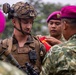 A U.S. Marine in full combat gear is receiving what appears to be a patch or insignia from a foreign military soldier wearing a distinctive magenta/pink beret. The scene suggests a joint military exercise or ceremony involving U.S. Marines and what appears to be Indonesian Marine Corps (Korps Marinir) personnel, identifiable by their characteristic magenta berets and camouflage uniforms. This type of exchange is common during bilateral military training exercises.