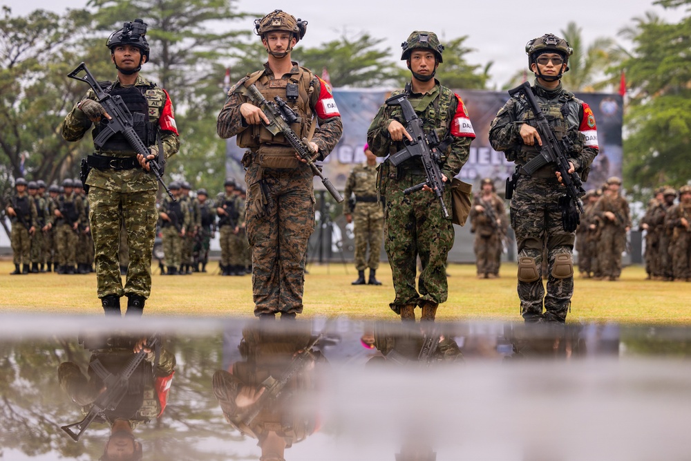Four armed soldiers from what appear to be different national militaries stand in formation on a grass field, each wearing distinct camouflage uniforms and carrying assault rifles. They all wear red and white armbands, suggesting a joint multinational military exercise or ceremony. Their reflections are visible in a puddle of water in the foreground, and rows of additional troops are visible in the background.