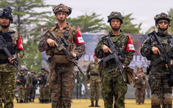 Four armed soldiers from what appear to be different national militaries stand in formation on a grass field, each wearing distinct camouflage uniforms and carrying assault rifles. They all wear red and white armbands, suggesting a joint multinational military exercise or ceremony. Their reflections are visible in a puddle of water in the foreground, and rows of additional troops are visible in the background.