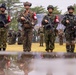 Four armed soldiers from what appear to be different national militaries stand in formation on a grass field, each wearing distinct camouflage uniforms and carrying assault rifles. They all wear red and white armbands, suggesting a joint multinational military exercise or ceremony. Their reflections are visible in a puddle of water in the foreground, and rows of additional troops are visible in the background.