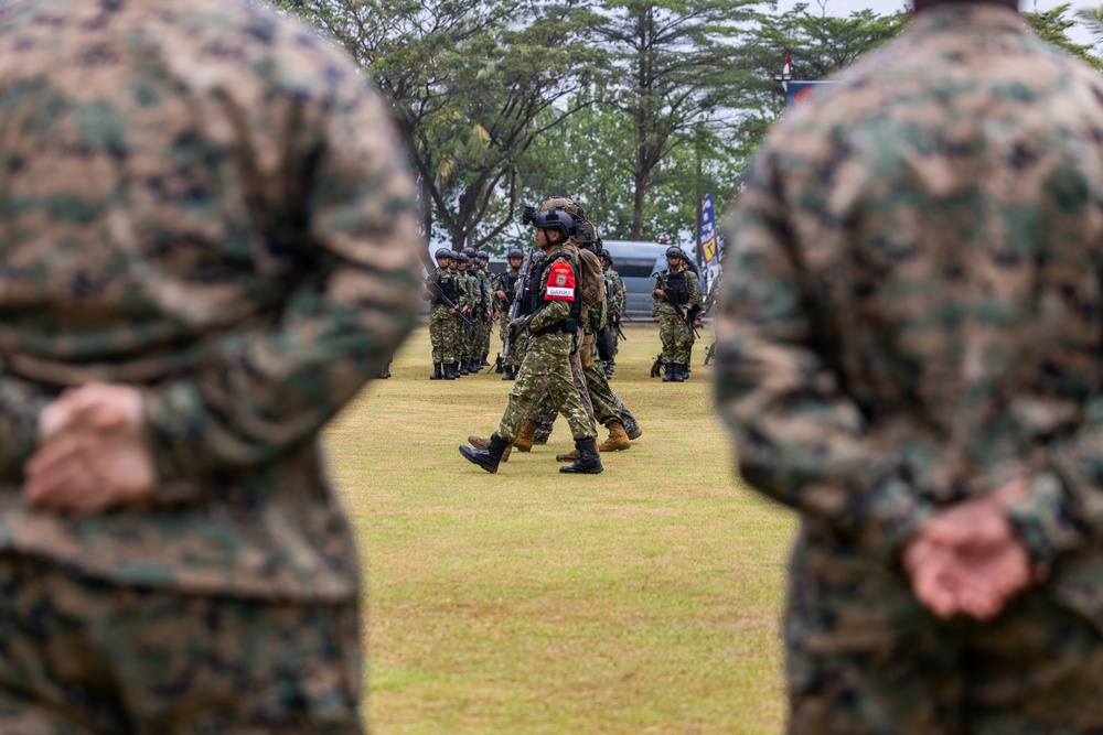 A military exercise or demonstration is taking place on an open grass field, with soldiers from at least two different nations participating. Two soldiers in MARPAT (US Marine Corps digital camouflage) are visible in the foreground with hands clasped behind their backs in an 'at ease' stance, observing personnel in a different camouflage pattern marching in the middle ground. The central figure wears a red patch on their arm and carries full combat gear including a helmet-mounted device.