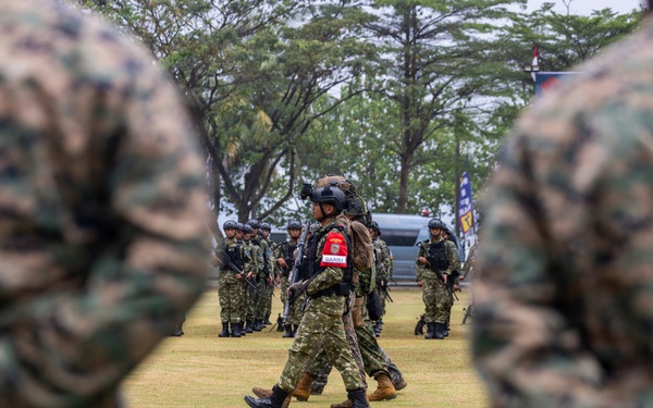 A military exercise or demonstration is taking place on an open grass field, with soldiers from at least two different nations participating. Two soldiers in MARPAT (US Marine Corps digital camouflage) are visible in the foreground with hands clasped behind their backs in an 'at ease' stance, observing personnel in a different camouflage pattern marching in the middle ground. The central figure wears a red patch on their arm and carries full combat gear including a helmet-mounted device.