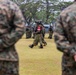 A military exercise or demonstration is taking place on an open grass field, with soldiers from at least two different nations participating. Two soldiers in MARPAT (US Marine Corps digital camouflage) are visible in the foreground with hands clasped behind their backs in an 'at ease' stance, observing personnel in a different camouflage pattern marching in the middle ground. The central figure wears a red patch on their arm and carries full combat gear including a helmet-mounted device.