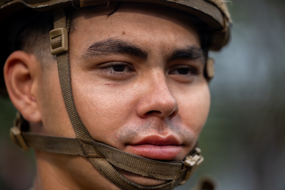 A close-up portrait of a young male soldier wearing a military helmet with a tan/olive chin strap secured under his jaw. The image is a tight facial shot showing intense, focused expression with the subject gazing slightly off-camera. The background is blurred, suggesting an outdoor environment with muted green tones.