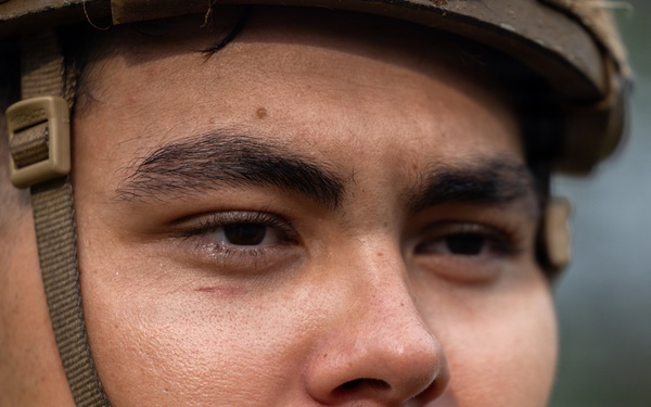A close-up portrait of a young male soldier wearing a military helmet with a tan/olive chin strap secured under his jaw. The image is a tight facial shot showing intense, focused expression with the subject gazing slightly off-camera. The background is blurred, suggesting an outdoor environment with muted green tones.
