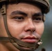 A close-up portrait of a young male soldier wearing a military helmet with a tan/olive chin strap secured under his jaw. The image is a tight facial shot showing intense, focused expression with the subject gazing slightly off-camera. The background is blurred, suggesting an outdoor environment with muted green tones.