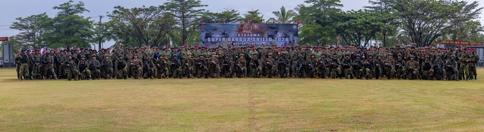 A large group of military personnel from multiple nations pose for a formal group photograph at the 'Super Garuda Shield 2026' joint military exercise held at Marinir Lampung. The participants are arranged in rows on a grass field, wearing various military uniforms and combat gear. A large banner identifying the exercise is prominently displayed behind the assembled troops.